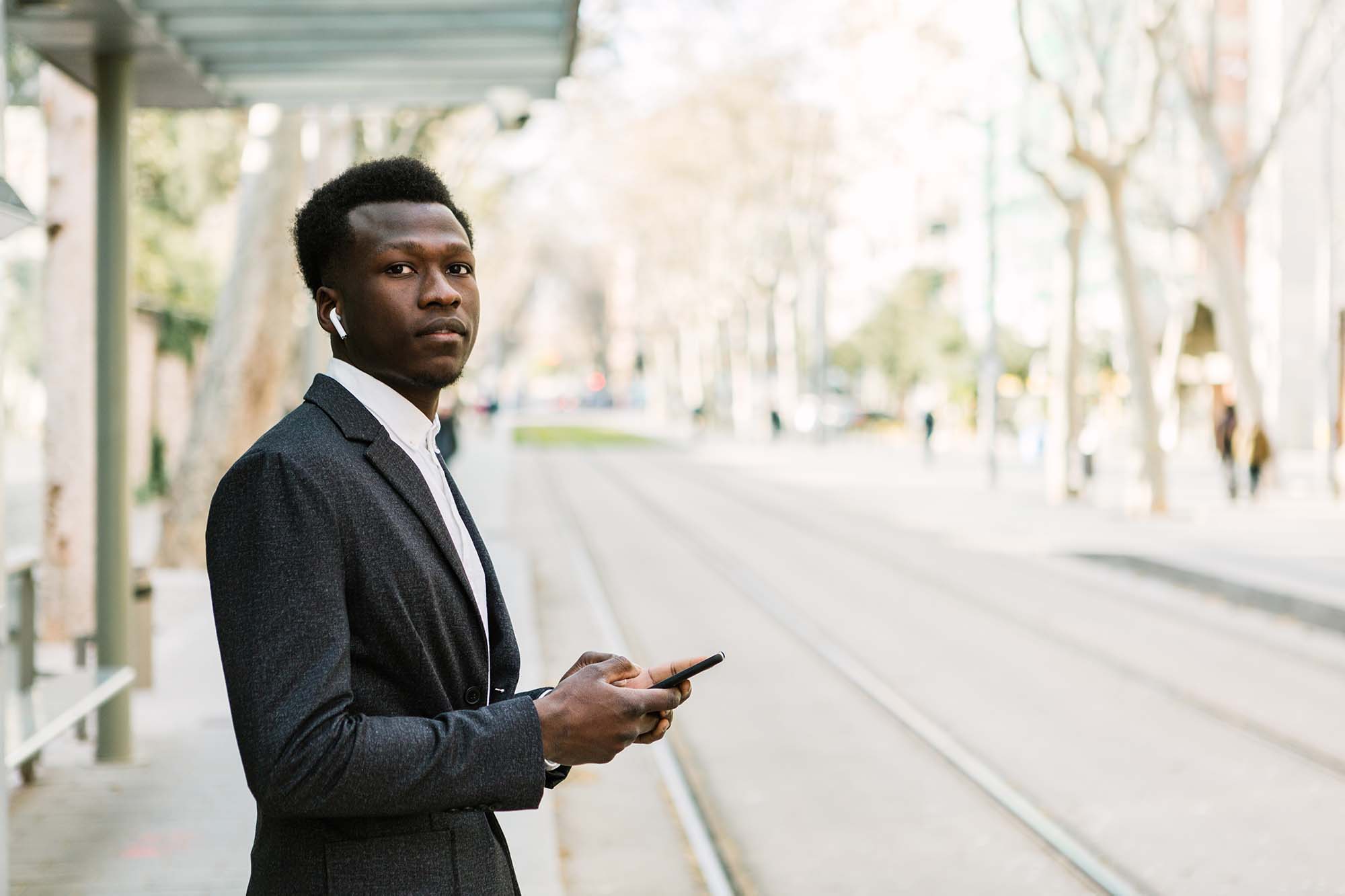 Young black businessman listening to music with wireless headphones and using his phone while waiting for the train