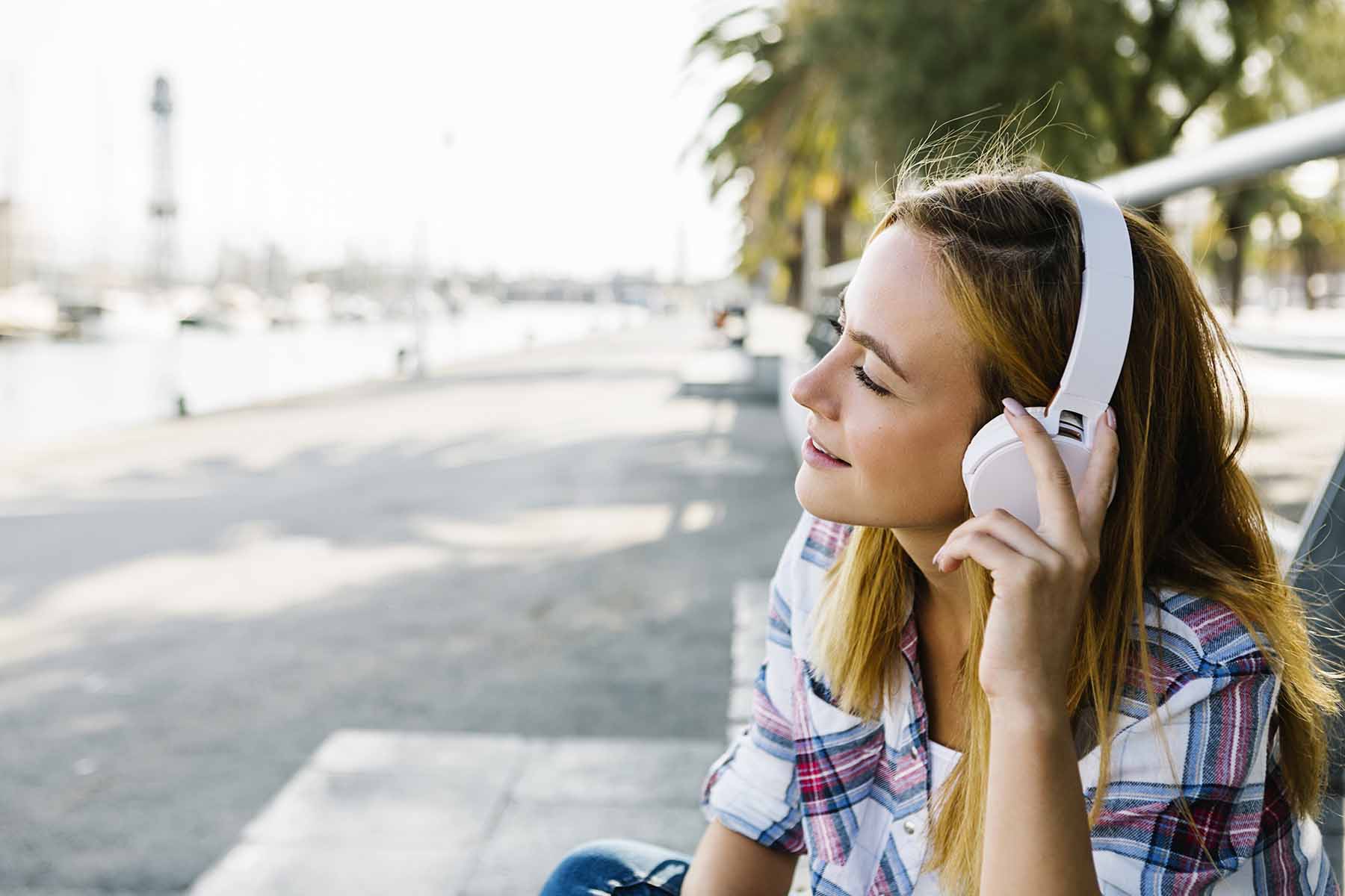 Young woman listening music through headphone sitting on footpath during sunny day