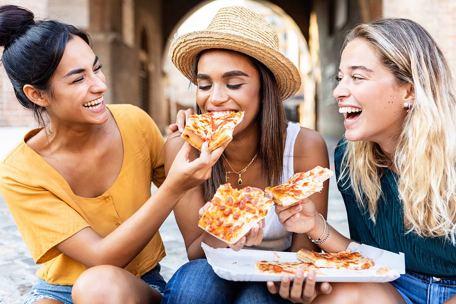 Three cheerful multiracial women eating pizza in the street